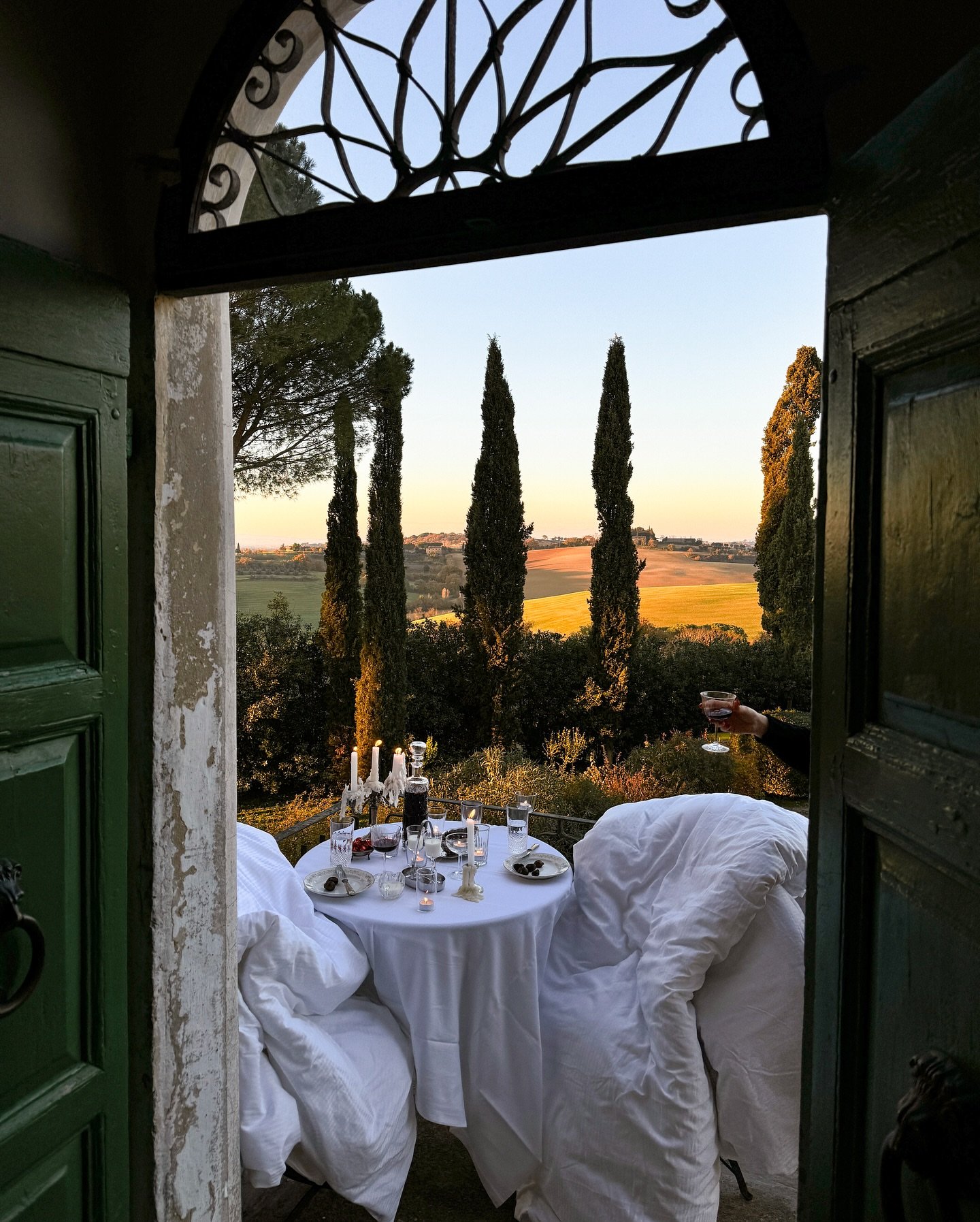 A candlelit wine dinner seen through green doors, Tuscany at dusk.