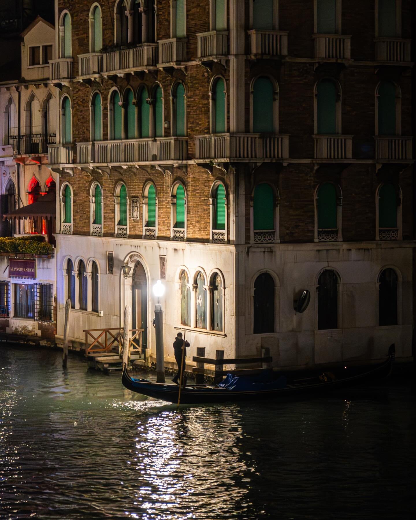 A gondolier passing beneath a lamplit palazzo after dark